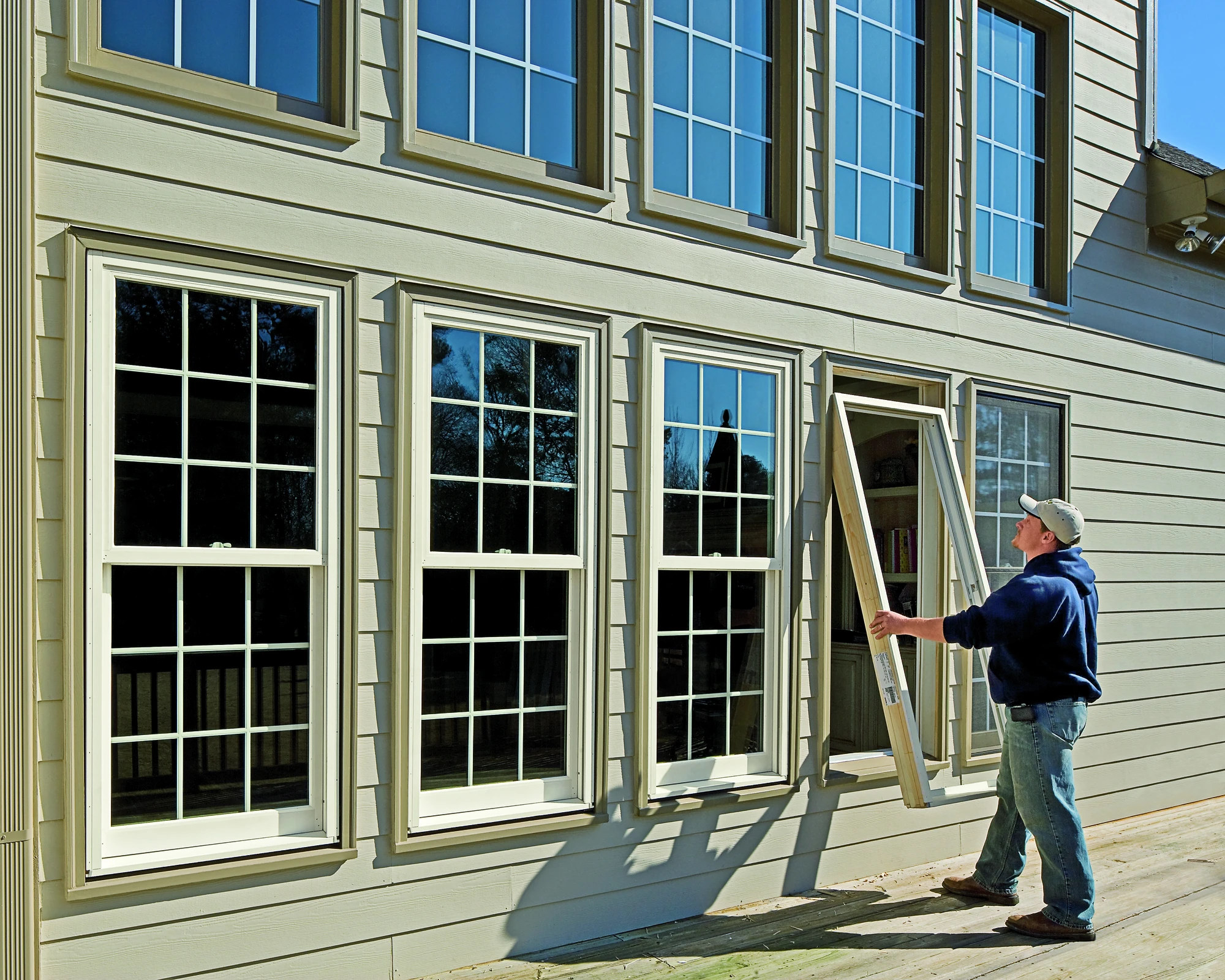 A worker installs a replacement window on the exterior of a beige two-story house with multiple white-trimmed windows