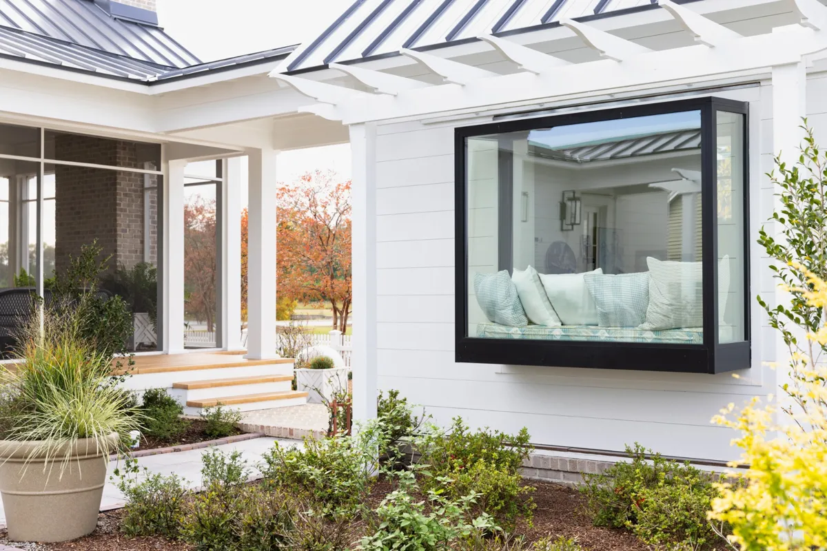 Modern white home exterior featuring a large black-framed box bay window with a cushioned seating nook, surrounded by landscaping and a covered porch