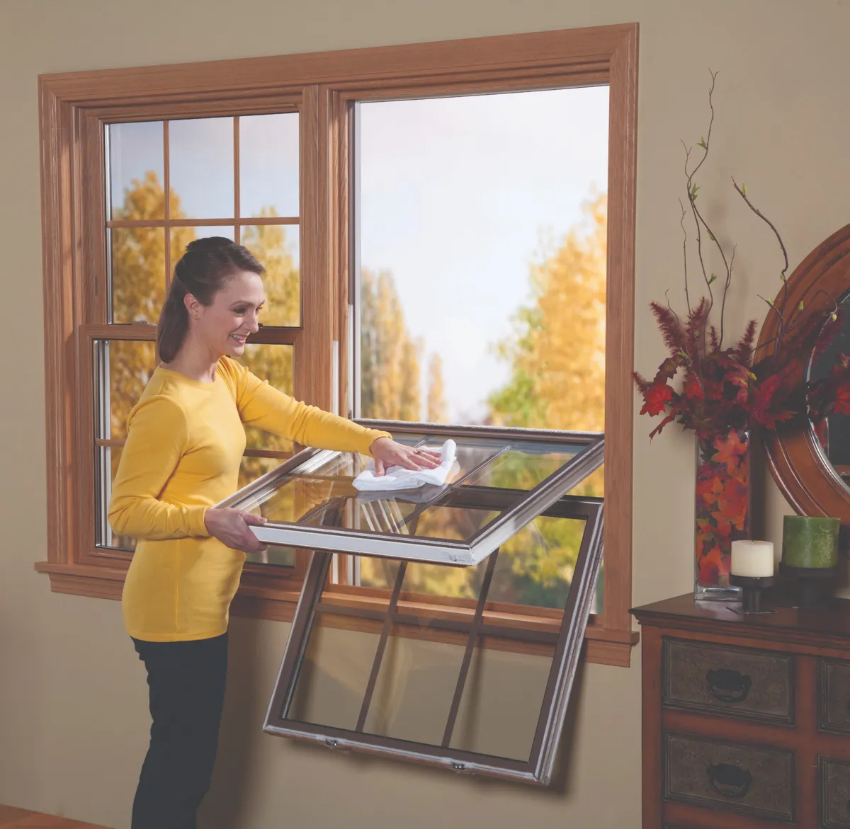 Woman in yellow shirt cleaning a tilt-in double-hung window with wood trim in a cozy, traditionally styled room with fall decor